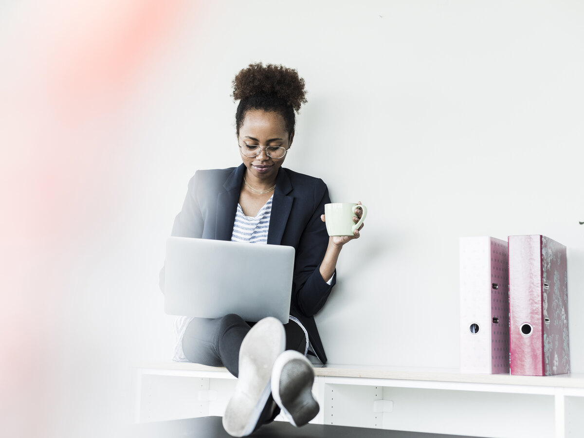 Une femme assise à un bureau clair, son ordinateur portable sur les genoux et un café à la main